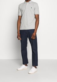 Harmaa polo-paita, jossa on kaulus, vihreä logo ja lyhyet hihat, yhdistettynä tummansinisiin farkkuihin ja valkoisiin tennareihin rentoa istuvuutta varten.