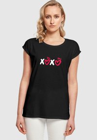 Svart t-skjorte med korte ermer som har hvit og rød "XOXO" grafikk med hjerter. Avslappet passform, rund hals, mykt stoff.
