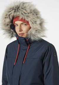 Navy parka with large faux fur hood, red drawstrings, and ribbed red beanie. Features front pockets and zipper closure. Smooth texture.