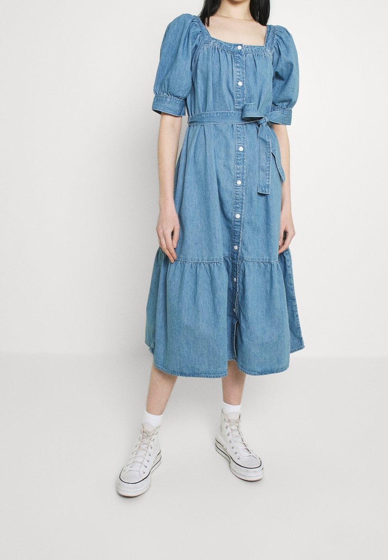Robe midi en jean bleu clair, manches ballon, encolure carrée, boutonnée devant, taille cintrée avec une ceinture, jupe à volants, portée avec des baskets blanches.