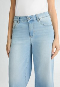 Vaqueros de mezclilla de tiro alto en azul claro con botón frontal, bolsillos y corte recto, combinados con una blusa blanca de canalé.