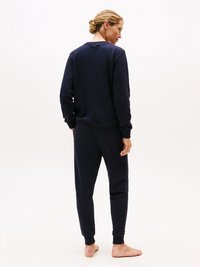 Kvinna barfota stående, vänd bort, iförd en mörk marinblå sweatshirt och matchande joggbyxor mot en ljus, enfärgad bakgrund.