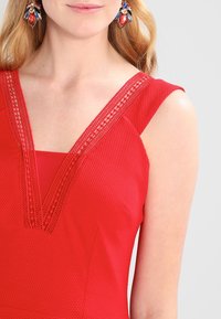 Robe sans manches rouge avec un tissu texturé et un empiècement en dentelle au niveau du col. Présente une forme en V prononcé et des lignes épurées, mettant en valeur les épaules.