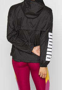 Svart lättviktsjacka med huva, som har elastiska ärmslut och en framträdande vit logotyp på vänster ärm. Slät textur och avslappnad design.