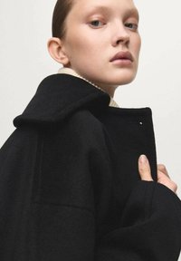 Abrigo negro de lana con cuello ancho, corte relajado, costuras visibles y tejido texturizado, llevado sobre un suéter de color claro.