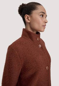 Cappotto di lana marrone con un colletto alto, presenta due bottoni marroni e un tessuto texturizzato. Vista laterale che mette in mostra i dettagli del colletto e dei bottoni.