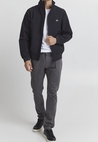 Giacca bomber nera con colletto alto, chiusura con zip, polsini elastici, abbinata a pantaloni grigi e sneakers nere su uno sfondo neutro.