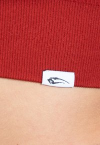 Tejido de ribetes rojos con una etiqueta blanca que presenta un logo gráfico negro. Enfoque en la textura y el patrón, con un acabado suave y un ligero estiramiento.