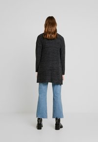 Lång svart cardigan i texturerad stickning, kombinerad med ljusblå utsvängda jeans och svarta ankelboots. Designen har en avslappnad passform.