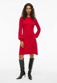 Rotes langärmliges Kleid mit geradem Schnitt und Ballonärmeln, aus weichem Stoff. Kombiniert mit knielangen schwarzen Stiefeln mit Absätzen.