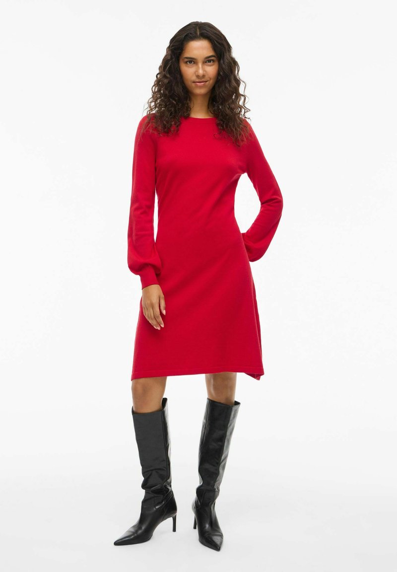 Rotes langärmliges Kleid mit geradem Schnitt und Ballonärmeln, aus weichem Stoff. Kombiniert mit knielangen schwarzen Stiefeln mit Absätzen.