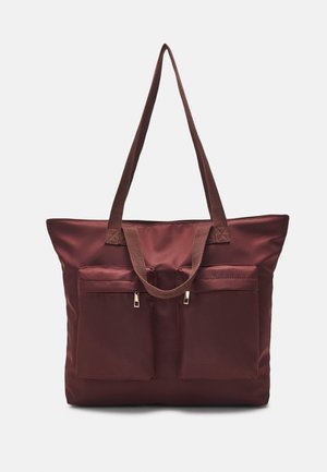 Duża ciemnobrązowa torba na ramię z dwoma przednimi kieszeniami zapinanymi na suwak oraz długimi paskami na ramię na jednolitym jasnym tle.