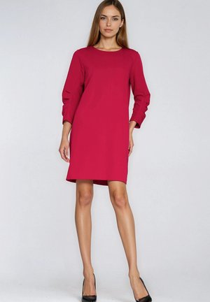 Kobieta ubrana w czerwoną sukienkę do kolan z długimi rękawami z marszczeniami, stojąca na tle jednolitego jasnoszarego tła.