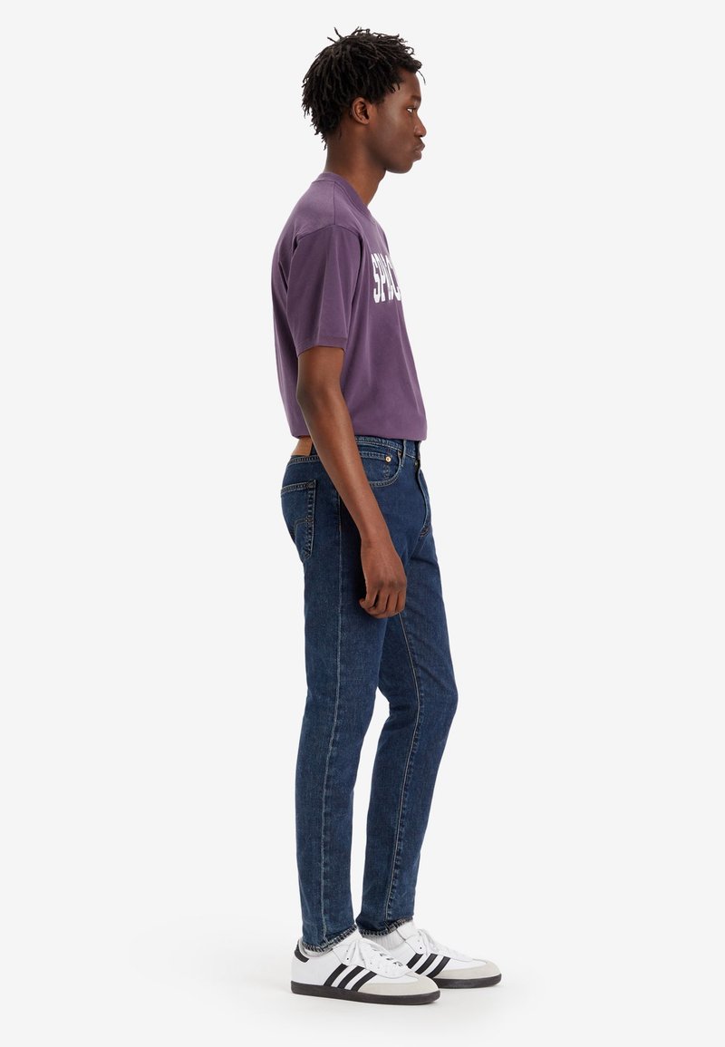 Lila T-Shirt mit kurzen Ärmeln und weißem Text, kombiniert mit dunkelblauen Slim-Fit-Jeans und schwarz-weißen Sneakers, von der Seite betrachtet.