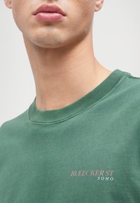 Zaļš kokvilnas kreklis ar īsām piedurknēm un apaļu kakla izgriezumu, ar rozā krāsā izdrukātu uzrakstu "BLEECKER ST SOHO" uz krūtīm, gluda tekstūra un minimālisma dizains.