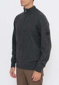 Uomo che indossa un maglione grigio scuro con mezza zip, polsini e orlo a coste, abbinato a pantaloni marroni, su sfondo bianco semplice.