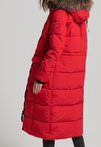 Rode pufferjas met een met bont afgewerkte capuchon en ritssluiting. Kenmerken zijn het horizontale quiltontwerp, twee voorzakken en een logo-patch op de mouw.