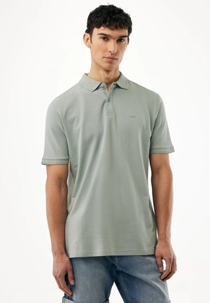 Junger Mann mit kurzen dunklen Haaren, der ein hellgrünes Poloshirt und blaue Jeans trägt, vor einem einfarbigen weißen Hintergrund stehend.