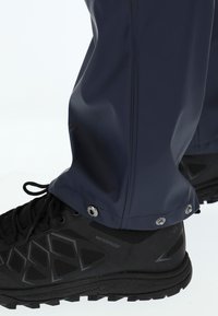 Vedenpitävät tummansiniset housut, joissa on sileä ja kiiltävä pinta sekä nilkoissa painonappikiinnitykset. Yhdistetty mustiin, kestävistä materiaaleista valmistettuihin vaelluskengiin.