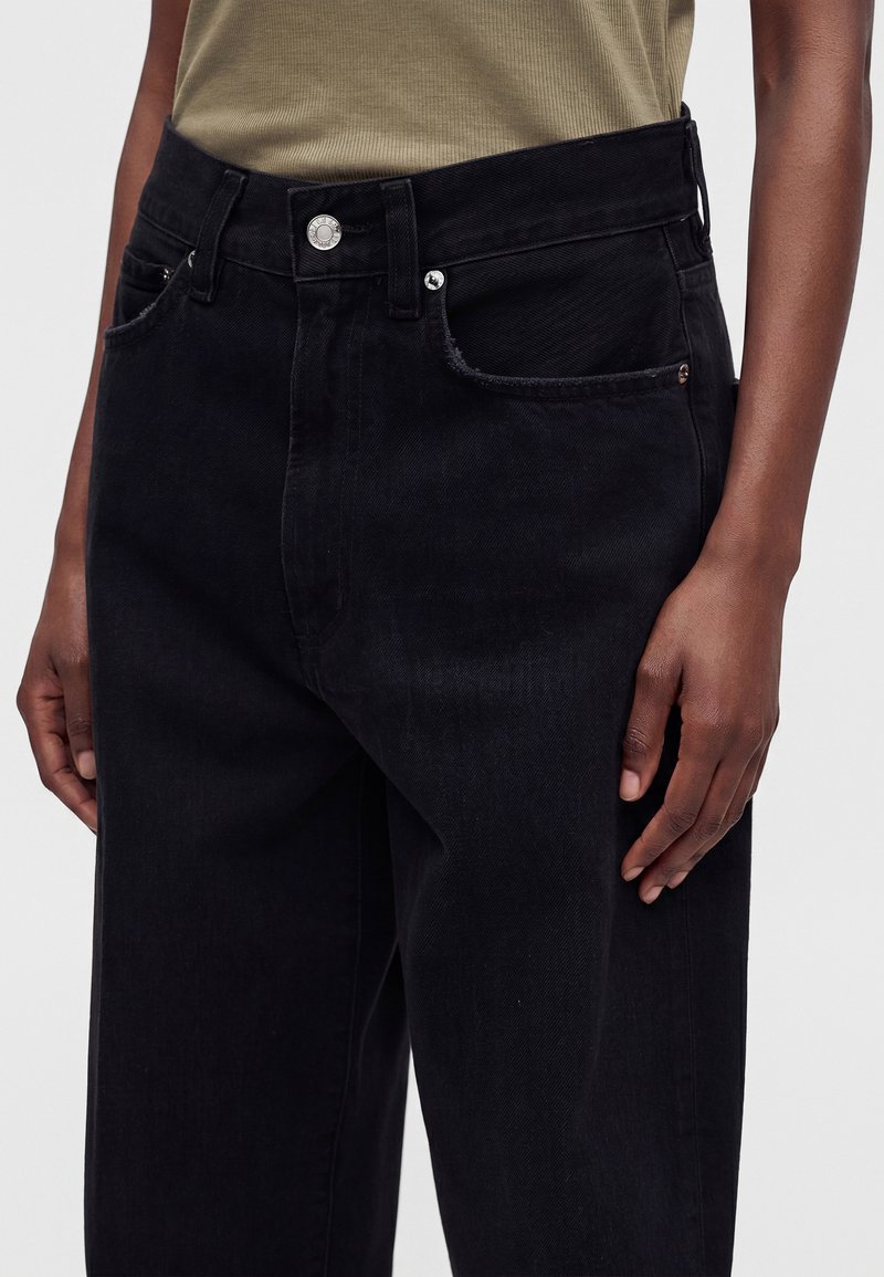 Personne portant un jean noir ample taille haute et un top sans manches côtelé vert olive, debout, les bras détendus.