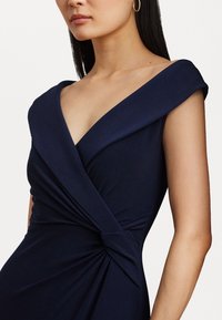 Dunkelblaues Kleid mit tiefem V-Ausschnitt und drapiertem Schulterdesign. Mit einer verdrehten Detail am Taillenbereich für eine figurbetonte Silhouette. Glatte Stofftextur.