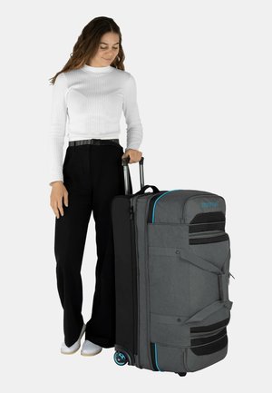 Graue Rolltasche mit schwarzen Akzenten, zwei Rädern, Seitengriffen, Reißverschlüssen und türkisen Trims, die von einer Person in lässiger Kleidung getragen wird.