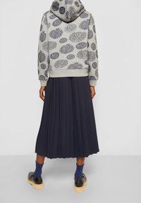 Persona che indossa un felpa grigia con motivi circolari, una gonna midi plissettata blu navy, calzini blu e scarpe nere con suole beige, vista da dietro.