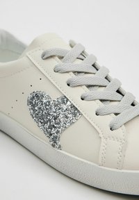 Zapatilla de cuero blanca con un acento de corazón de brillo plateado, cordones grises, suela texturizada y perforaciones en el costado para transpirabilidad.