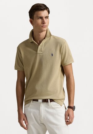 Polo beige en tissu texturé, doté d'une patte de boutonnage à trois boutons et d'un logo bleu sur la poitrine gauche, associé à un jean de couleur claire.