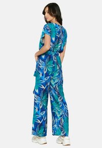 Gėlėtas kombinezonas mėlynos ir žalios spalvų su plačiomis kelnėmis, trumpomis rankovėmis ir per visą liemenį sutrauktu juosmeniu. Yra botaninis raštas ir laisvas siluetas.