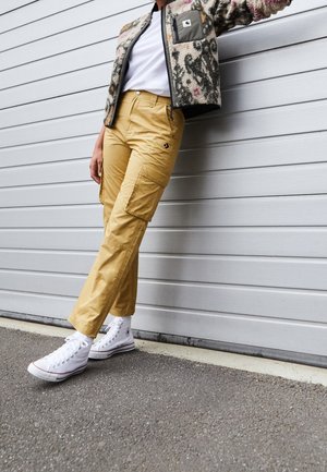 Person wearing beige cargo pants, white high-top sneakers, white shirt, and patterned fleece jacket leaning against a light gray wall.