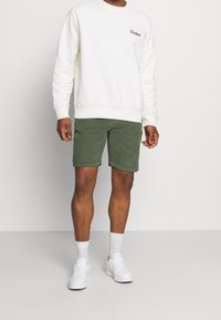 Witte sweatshirt met "Dickies" logo, groene shorts en witte sneakers. Het sweatshirt heeft een textuurafwerking en de shorts hebben een klassieke fit en zoom.