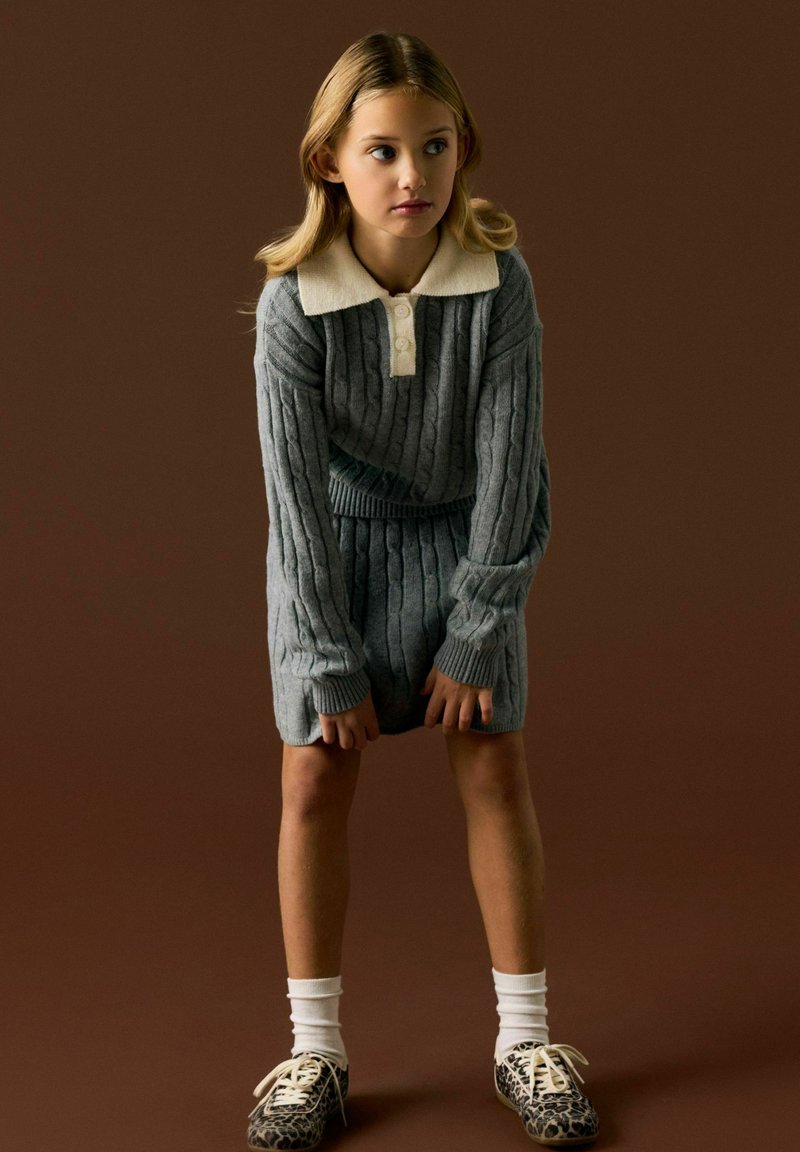Fille portant un pull en maille torsadée gris et un short, col blanc, chaussettes blanches et chaussures imprimé léopard, debout devant un fond marron.