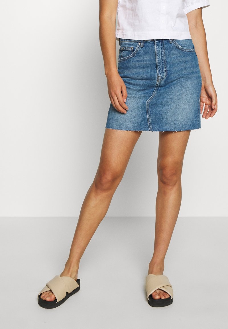 Džínová mini sukně ve středně modré barvě s třepeným lemem, s předními kapsami a těsným střihem. V páru s béžovými nazouvacími sandály.
