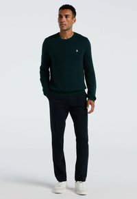 Dunkelgrüner Strickpullover mit rundem Halsausschnitt, gerippten Bündchen und Saum; kombiniert mit navyblauen Hosen und weißen Sneakern. Enthält kleines Logo auf der Brust.