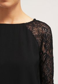 Top nero con maniche corte in pizzo nero caratterizzate da un motivo floreale, scollo rotondo e corpino liscio. I dettagli sono semplici ed eleganti.