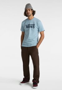 Ljust blå t-shirt med en mörkblå "VANS" logotyp, tillsammans med bruna byxor och vinröda sneakers. Modellen bär en grå stickad mössa.