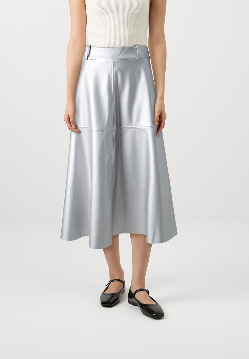 Femme portant une jupe midi argentée avec passants de ceinture, associée à un haut blanc sans manches et des chaussures Mary Jane noires.