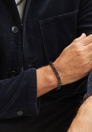 Zwarte metalen kettingarmband met in elkaar grijpende schakels, gedragen om een pols, vergezeld van een marineblauwe corduroy shirt met knoopdetails en een zak.