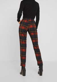 Person klädd i högmidjade rutiga byxor i röd, grön och marinblått tillsammans med en svart figurnära långärmad topp och svarta klackstövlar, stående med händerna i fickorna.