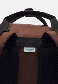 Hnědý batoh s černými síťovanými stranami, který má pevnou horní rukojeť a štítek s nápisem "Vegatex™." Texturovaný povrch z odolných materiálů.