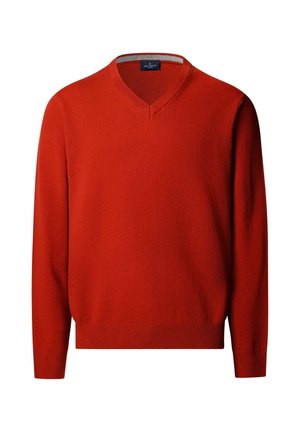 Hombre de pie con las manos en los bolsillos, vistiendo un suéter rojo de cuello en V, camisa blanca, pantalones marrones y zapatos negros sobre un fondo claro y liso.