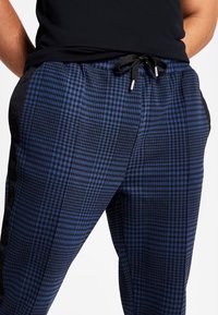 Námořnické modré kostkované kalhoty s texturovanou tkaninou, vyznačující se pasem na šňůrku a černými bočními akcenty. Navrženo pro pohodlí a styl.