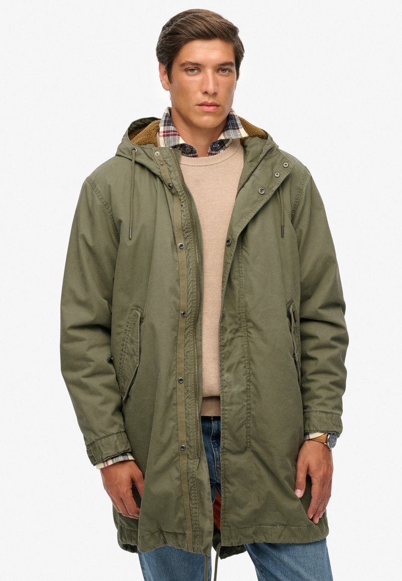 Abrigo largo verde oliva con capucha, cierre de botones a presión y cremallera, forro texturizado, bolsillos laterales, llevado sobre un suéter beige y una camisa de cuadros.