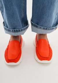 Červené kožené boty na slip-on s bílými gumovými podrážkami a strukturovanými švy. Nosené s ohrnutými modrými džínami, což vytváří neformální styl.