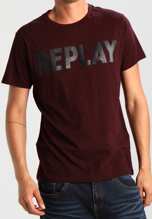 Mand iført en mørk vinrød kortærmet T-shirt med skinnende sort "REPLAY" tekst hen over brystet og mørkeblå jeans.