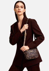 Borsa a tracolla marrone con motivo, con cerniera dorata e tracolla regolabile caratterizzata da dettagli con logo; tenuta da una modella che indossa un blazer scuro.