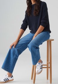 Pull en maille côtelée marine, jean à jambes larges bleu clair et baskets blanches avec des rayures marines, assis sur un tabouret en bois devant un fond clair.