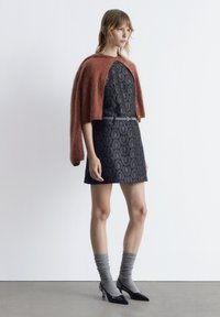 Schwarzes gemustertes Kleid mit einer figurbetonten Silhouette, kombiniert mit einem übergroßen strukturierten schokoladenbraunen Schal, grauen gerippten Socken und schwarzen spitzen Absätzen.