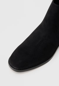 Čierne semišové kotníkové topánky s špicatou špičkou a hladkou textúrou. Obsahujú úzku čiernu podrážku a jemné švy okolo horného okraja.